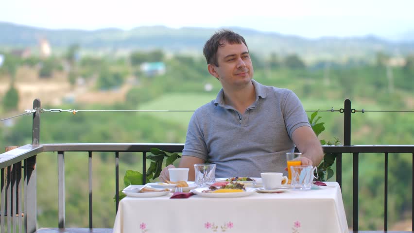 Man enjoying healthy breakfast with juice on balcony overlooking picturesque countryside. Relaxed dining experience in scenic outdoor setting with natural landscapes in background. Tranquil morning