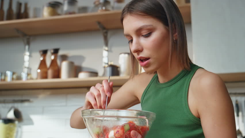 Girl eating vegetables salad in apartment kitchen closeup. Serious young woman enjoy healthy meal sitting contemporary cuisine. Attractive lady chewing vegetarian food for breakfast early morning.