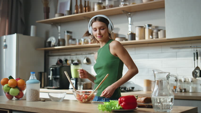 Earphones lady making salad in cozy kitchen dancing to music. Happy woman in wireless headphones preparing healthy meal in contemporary cuisine. Smiling girl mixing fresh ingredients at home counter.