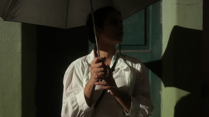 Woman in vintage dress holding umbrella, standing by green wall in sunlight