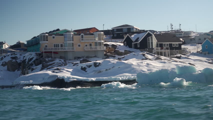 POV shot from a moving boat showcasing colorful homes and small icebergs near a rugged, snowy shore in Ilulissat, Greenland