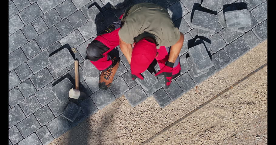A person skillfully lays down paving stones in a residential driveway, ensuring each piece fits perfectly while working under the sun.