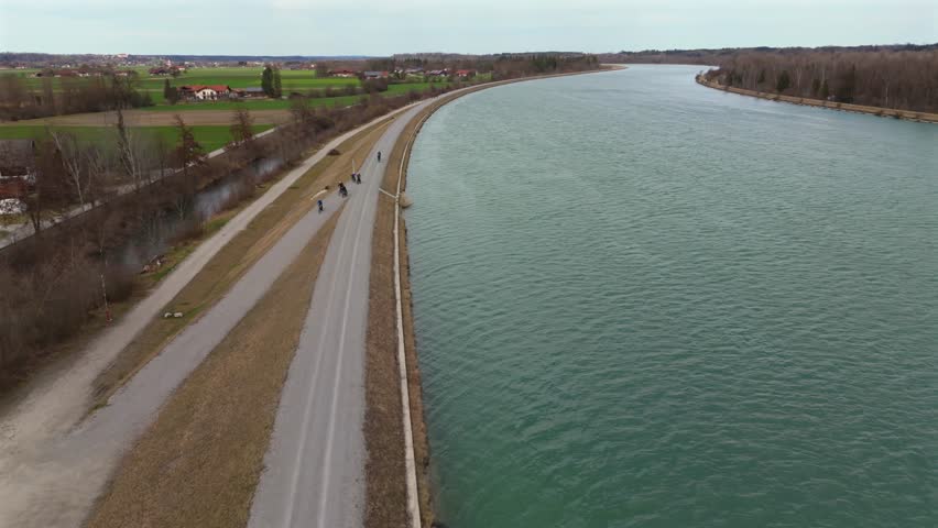 Cyclists ride on an embankment bicycle path along River Innnear Rosenheim, Bavaria, Germany. Am Innradweg bei Rosenheim. Gravel bike path along a wide river in Deutschland. Cycling in upper Bayern. 