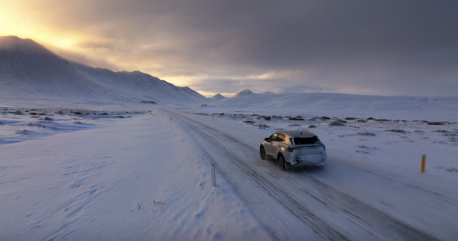 Aerial shot of a vehicle driving along a snow-covered road at dusk, with vibrant sunset hues casting a beautiful glow over the mountain landscape