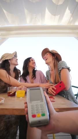Vertical young group of only women paying the bill with a nfc cellphone in cocktail beach bar. Female holding a smart phone and giving a payment transaction to the cashier on vacations with friends