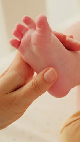 Mother holding in hands foot of newborn baby. Close up of woman gently stroking, touching tiny feet of baby boy. Family care, happy childhood and motherhood.