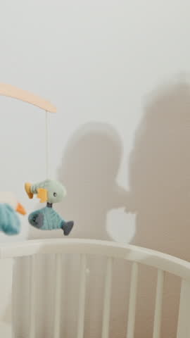 Silhouettes on white wall of the mother that playing with her little baby. Shadows near white cot with colourful crib mobile.