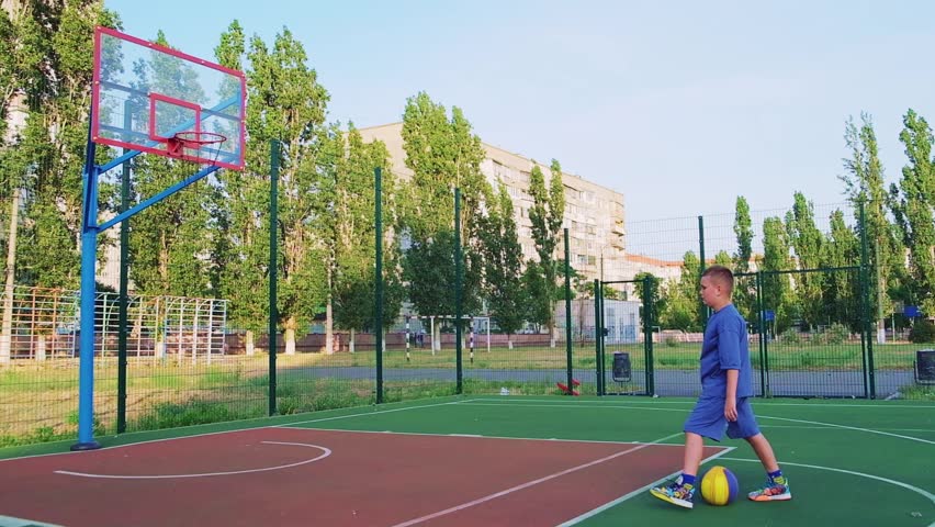Boy kid dressed in sports uniform playing basketball alone on street city sports playground. The concept of sports, active recreation, after school, workout and hobby