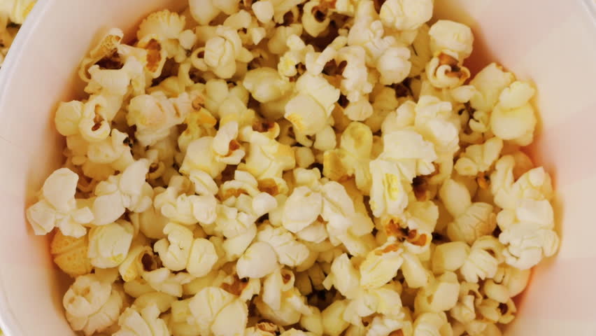 Popcorn grains falling into red and white striped bucket on background popcorn grains. Movie snack food, fast food cafe sweet snack. Slow motion. Top view.