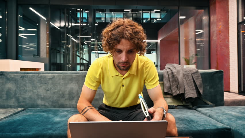 Man in yellow shirt standing on a couch and writing in a notebook and working on a computer