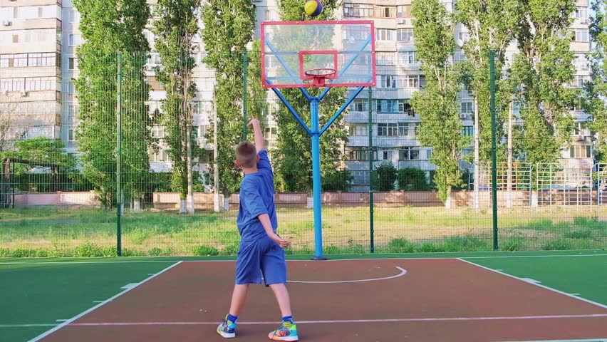 Boy kid dressed in sports uniform playing basketball alone on street city sports playground. The concept of sports, active recreation, after school, workout and hobby