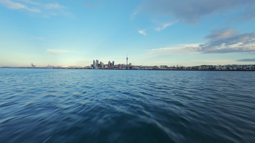 Aerial view flying low above the ocean toward the Auckland skyline, New Zealand. Fast aerial movement close to the water with the city, harbor, and Sky Tower visible on a sunny day.