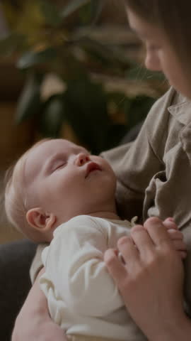 Vertical close-up of adorable Caucasian baby sleeping in arms of mother admiring her innocent little child, lovingly looking at cute face and holding tiny hand