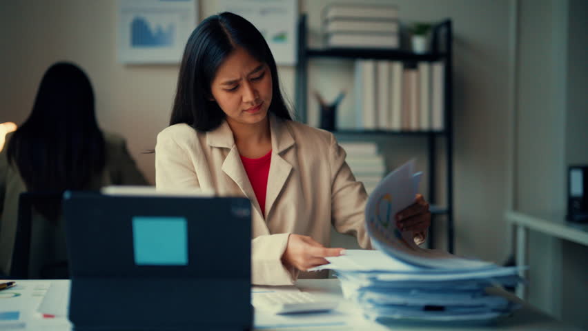 Focused businesswoman analyzing paperwork at her desk in a corporate office, surrounded by charts and office supplies. She exudes determination and professionalism in her work environment