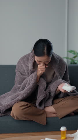 Young woman battling a cold, sitting on a sofa, blowing her nose, shivering under a blanket, looking miserable, and holding tissues