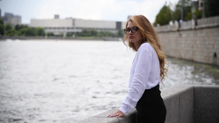 Portrait of beautiful blond long haired caucasian woman in eyeglasses and white shirt standing on city street on river embankment in a cloudy day. Soft focus. Copy space. People theme.