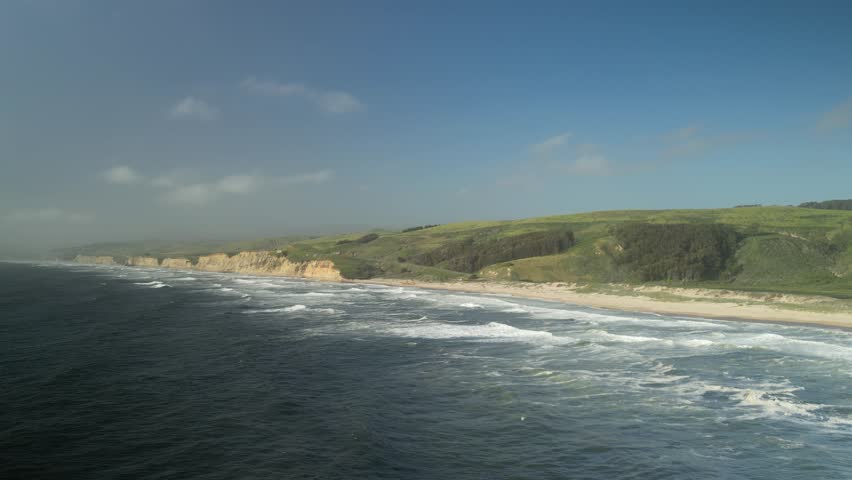 Aerial drone footage over Pescadero State Beach from the Peninsula of Northern California. The coastline features rugged cliffs, sandy shores, and crashing waves, all bathed in golden sunlight.