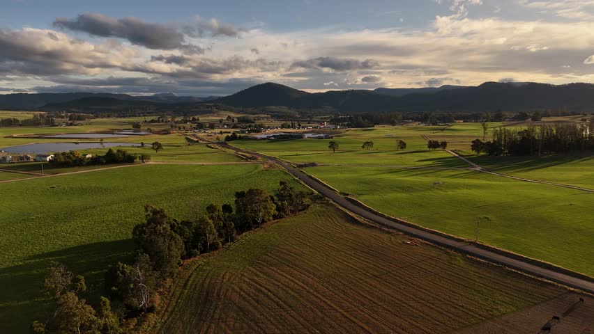 Magnificent Tasmanian landscape with green fields and main street at sunset. Mountain range in background. Small village with lake. Aerial wide shot.