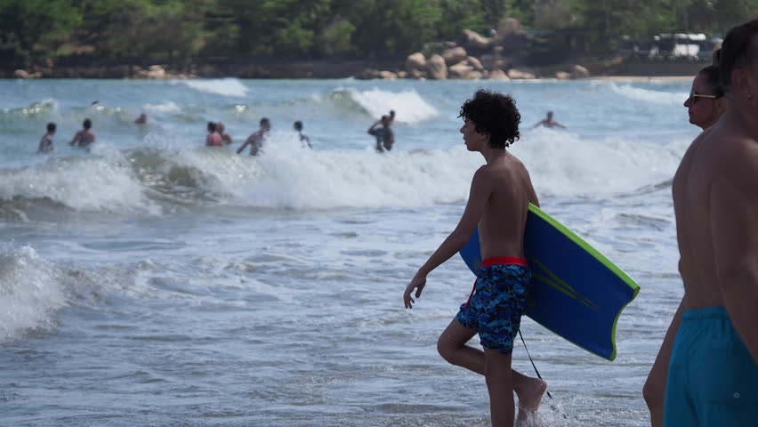 Nha Trang, Vietnam - March 25, 2024: A woman walks up to the boy with a small green surfboard, gesturing animatedly. The bustling summer beach is in the background.