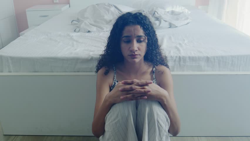 A distressed and anxious, curly-haired African woman sits on the bedroom floor, lost in painful thought. Concept of heartbreak, relationship turmoil, loneliness, and mental health issues
