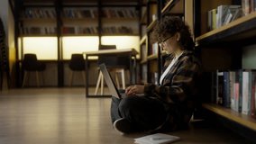A girl with curly hair wearing glasses sits on the floor near shelves with books and takes notes on a laptop in the library - Powered by Shutterstock - Get 15% off with code: PIKWIZARD15