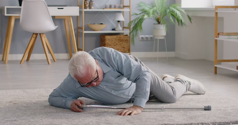 Elderly man experiences difficulties at home, trying to get up slowly after slipping and falling. Frail Caucasian pensioner with cane trying to stand up lying on floor in middle of living room.