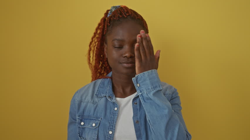 Confident african american woman standing, wearing denim shirt over a cheery yellow background, playfully covering one eye with hand, a smile plastered across her face