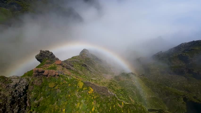 Rainbow over mountain ridge with clouds and sunlight. Aerial view from drone flying toward colorful rainbow in sky on Pico do Pico hiking trail, Madeira, Portugal.