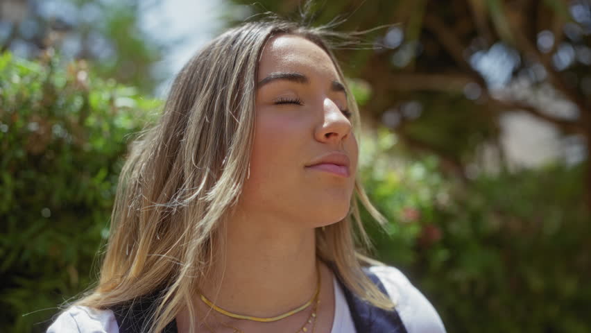 A serene young woman enjoys the sunny outdoors in a lush urban park, her blonde hair flowing in the breeze and eyes closed in contentment.