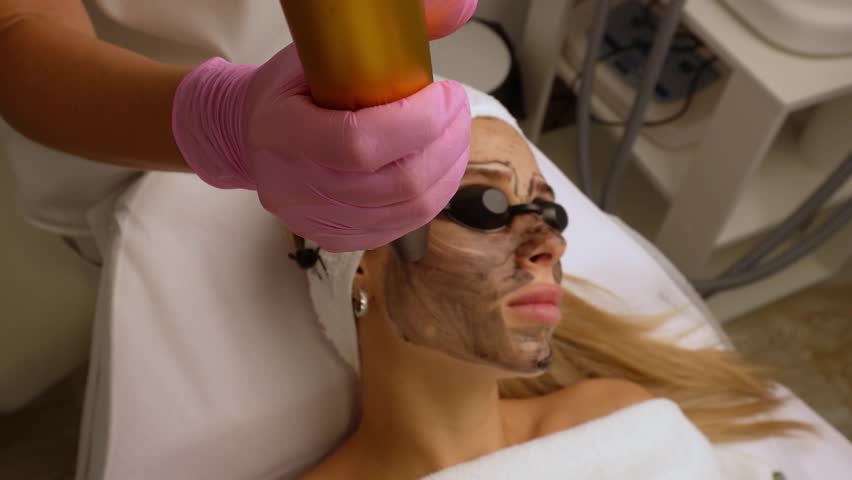 Beautician doing hardware carbon peeling to a young beautiful client in a headdress in goggles lying on a sofa in a beauty salon. Facial cleansing concept. Cosmetology services