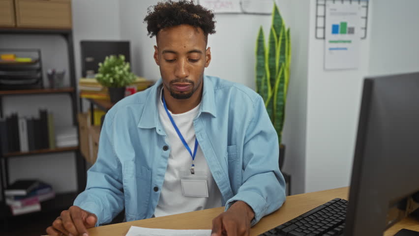 Young african american man with a beard working in an office, holding documents and wearing a blue lanyard, sitting at a desk with a computer and office supplies in the background.