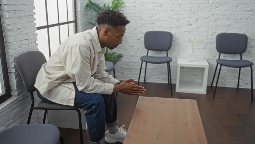 A pensive young man sitting in a modern waiting room, with chairs and a small table, wearing casual clothes and looking contemplative.