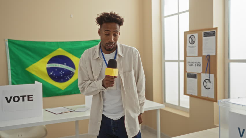 A young man with a beard stands indoors in an electoral college room in brazil, holding a microphone with a brazilian flag in the background.