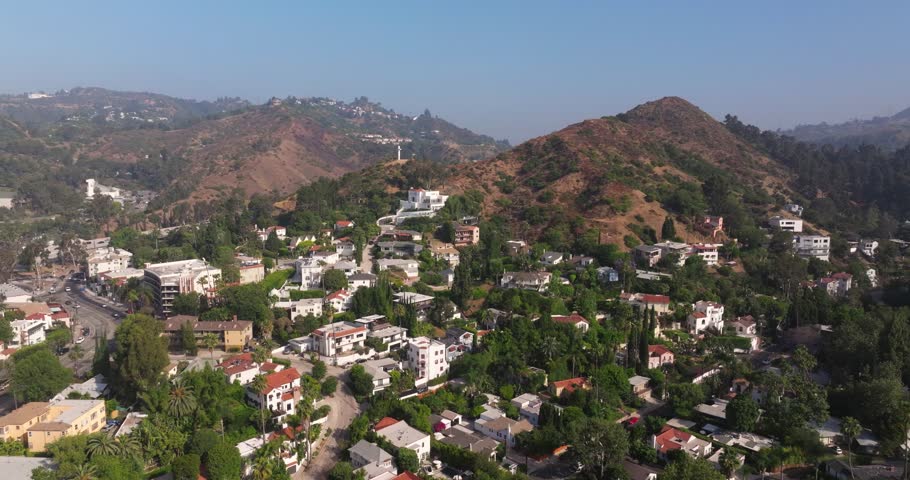 Panoramic aerial dolly over dense trees covering sidewalks up mountain side of hills in Los Angeles California with cross looking down