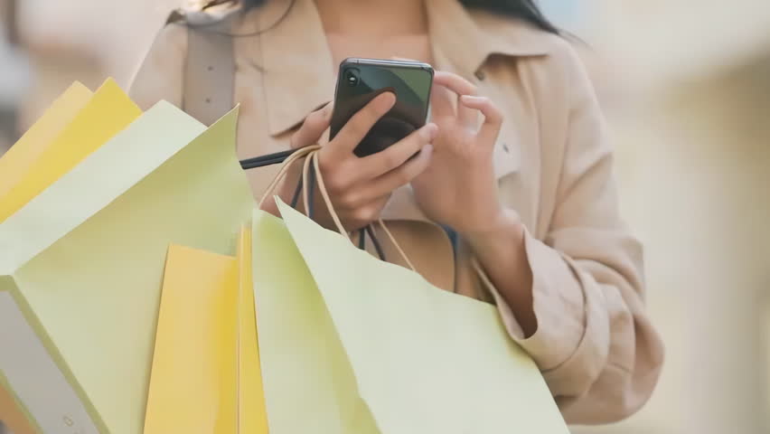 Asian happy woman young smiling girl in trendy stylish clothes with bright colorful shopping bags is using her smart phone while walking in the mall