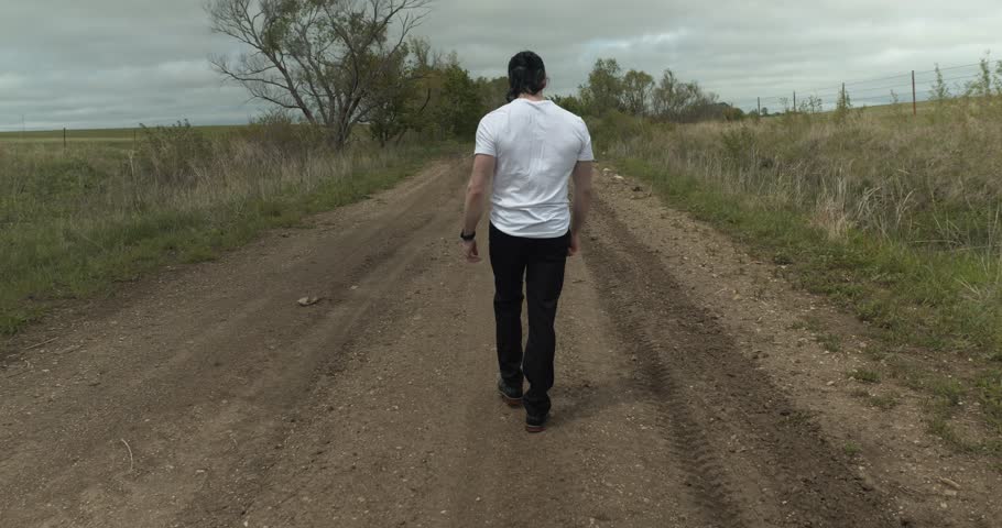 Man walking on dirt road by expansive fields. The overcast sky adds a moody atmosphere as he moves with determination, dressed in casual attire, embodying a sense of solitude and reflection.