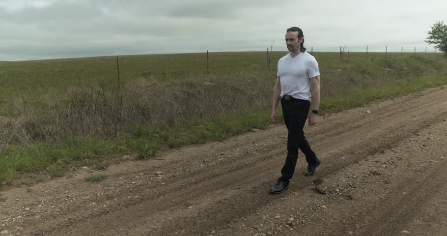 Man walking on dirt road by expansive fields. The overcast sky adds a moody atmosphere as he moves with determination, dressed in casual attire, embodying a sense of solitude and reflection.