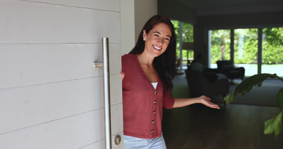 Cheerful young woman opens door, standing at doorway of modern apartment, warmly greets friend or neighbour with smile, extended hand showing welcoming gesture, invites invitee inside her cozy flat
