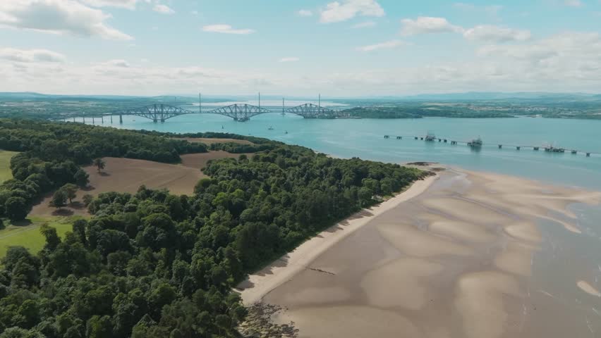 Aerial view of idyllic beach on Scottish coast. Scenic ocean views at low tide along shoreline of South Queensferry, Scotland. Lush green forest. Revealing famous bridges in background. United Kingdom