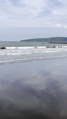 Portrait video footage from the beach with a view of the island and fishing boats above the waves of the sea that come to the white sand on a bright day with blue sky and white clouds