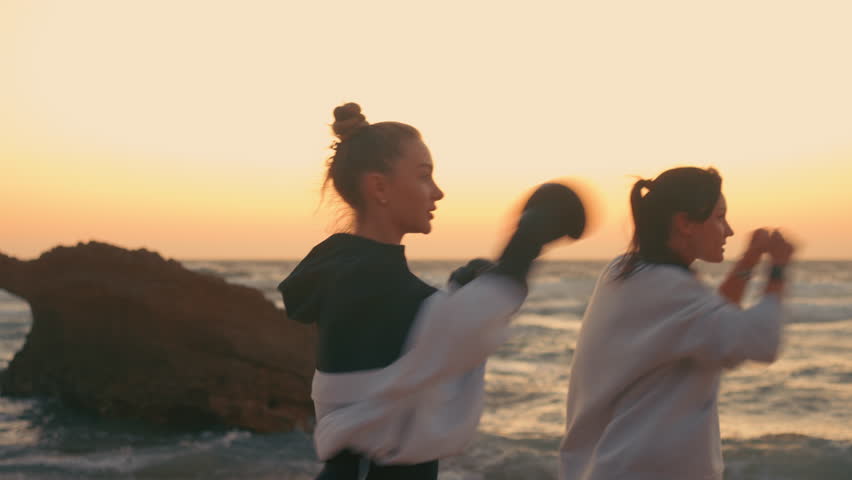 Determined sporty female fighters punching the air at beach during sunset. Side view of strong young women boxers exercising in nature. 