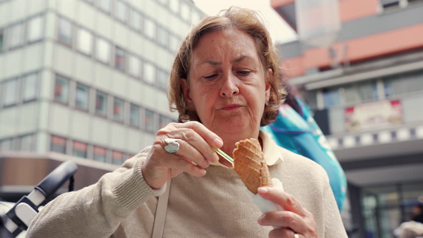 Older woman enjoying her ice cream cone outdoors, savoring each bite with a spoon, capturing a moment of contentment and sweet indulgence in a lively urban setting