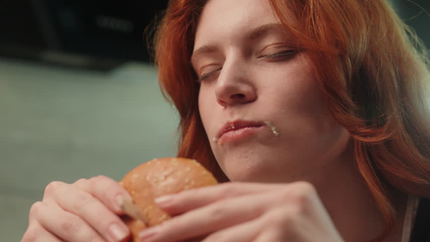 Close up hungry Caucasian pleased woman female client girl consumer with closed eyes chewing burger eat tasty hamburger bite cheeseburger enjoying taste indoor. Overeating healthcare unhealthy eating