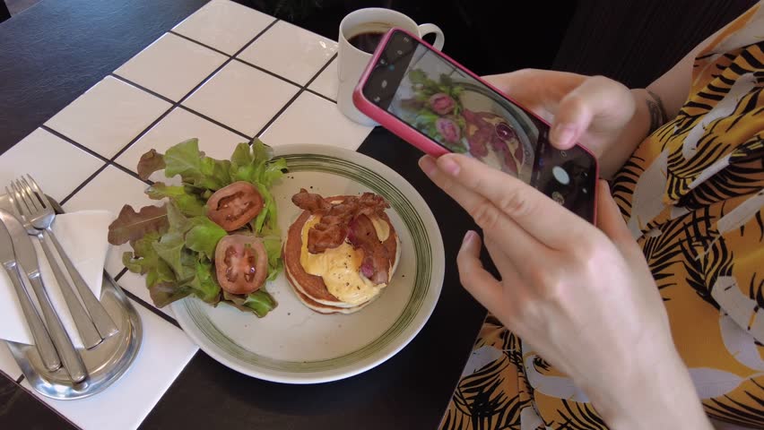 A person taking a video of their breakfast featuring pancakes, bacon, and fresh salad with a smartphone.