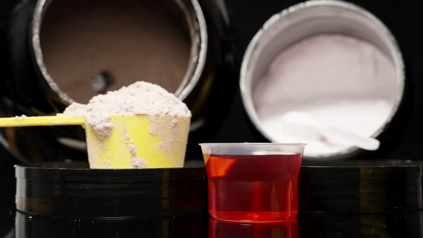 Slider Shot Across a Black Table as Red Sports Supplement Is Poured, with Protein Spoons Nearby. Pre-Workout Shake Preparation.