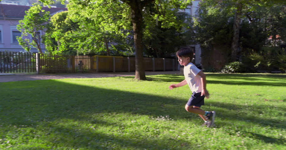 Young boy sprinting across a grassy park, with a determined expression, surrounded by trees and buildings, enjoying a sunny day outdoors