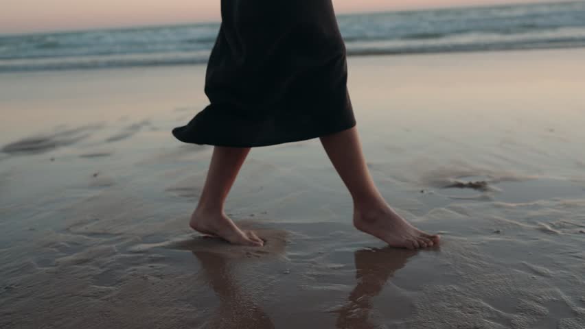A woman in a black dress walks barefoot on the wet sand of a beach at sunset. Her feet leave footprints in the sand.