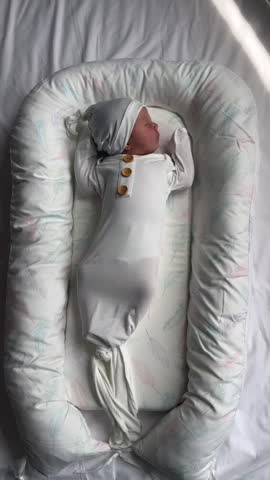 Newborn baby girl in a sleeping sack and a hat sleeping in a baby lounger. One week old baby napping in a baby nest. Vertical video