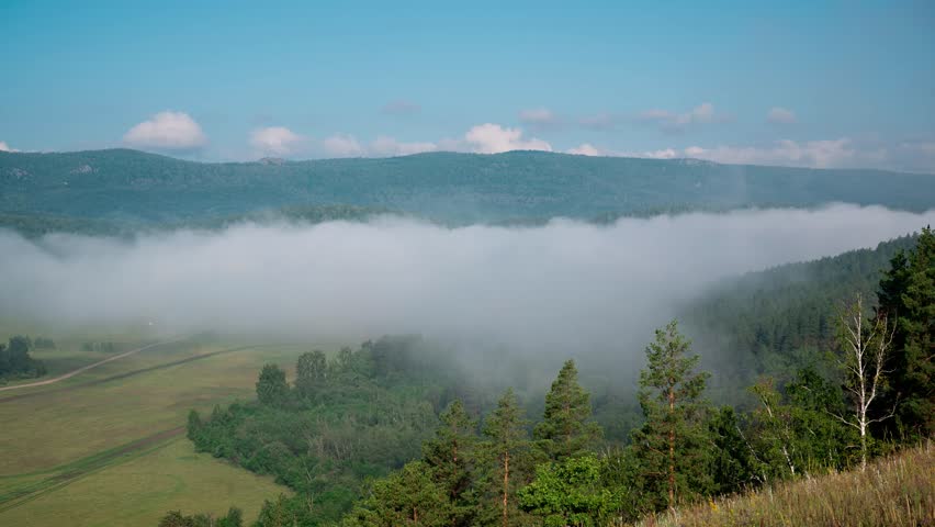 Fog rolling through a forested mountain valley on a clear morning, revealing a lush green landscape with distant mountains and fields. Ural mountains in Bashkortostan