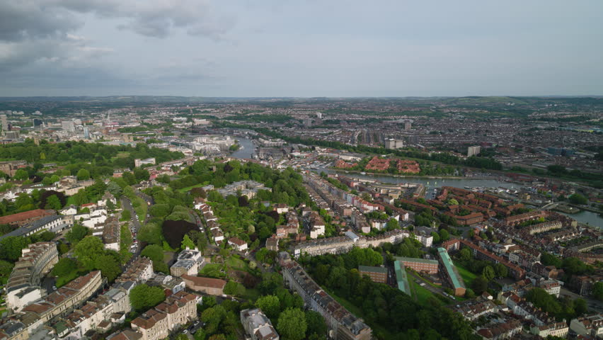 A stunning aerial view of Bristol, UK, on a sunny day, the cityscape shines with a mix of historic and modern architecture, surrounded by lush green parks and the winding River Avon